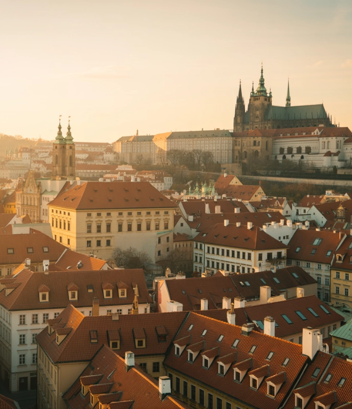 Panoramic view of Prague's historic center at sunrise, featuring the iconic red rooftops, St. Nicholas Church, and Prague Castle in the distance.