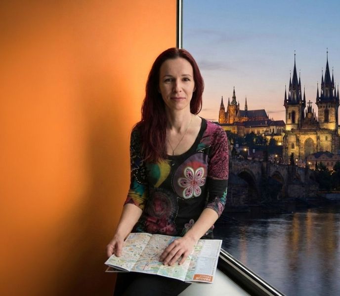 Portrait of Katerina Svobodova, founder of Prague Walker, smiling and holding an open map. A large window behind her reveals a view of the illuminated Charles Bridge and Prague landmarks at dusk, contrasting with an orange panel.